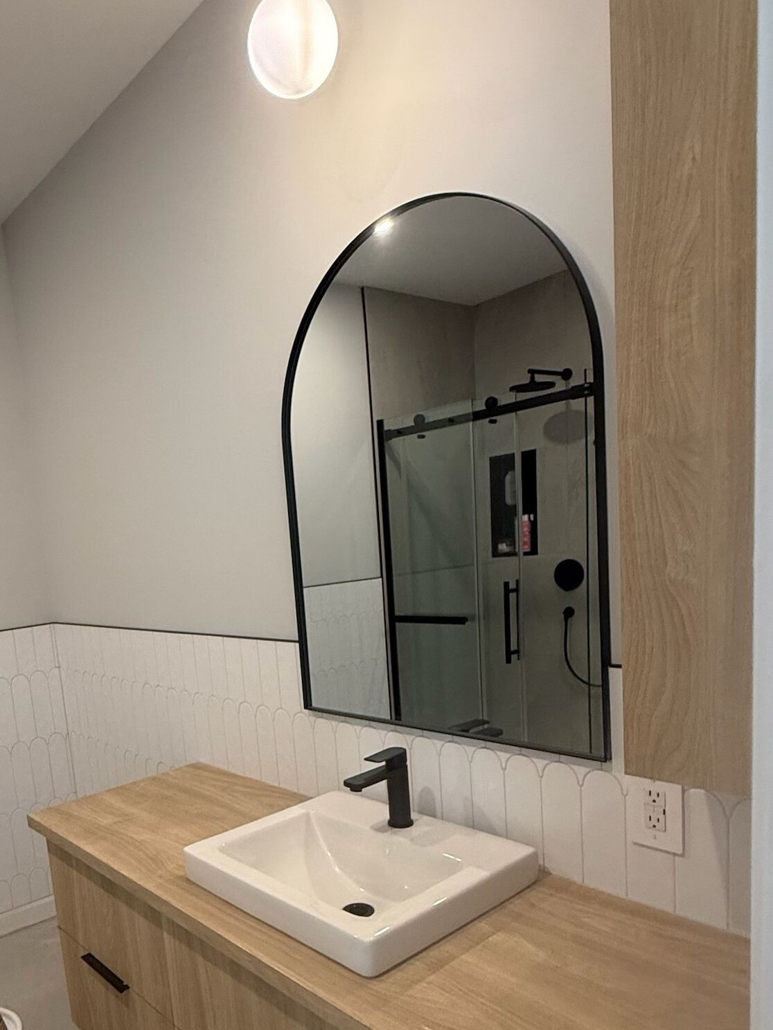 Salle de bain avec carreaux blancs en éventail à mi-mur, meuble en bois clair, miroir en arche et suspension